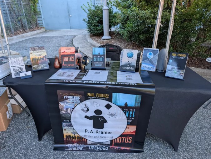 Promotional display for author P.A. Kramer, featuring various books and materials on a table, with a black tablecloth displaying a logo and website link.